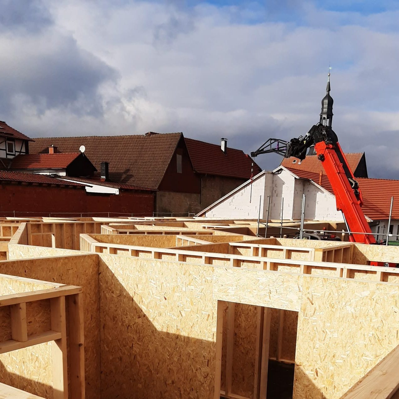 Holzrahmebau mit rotem Kran, im Hintergrund eine ländliche Umgebung und Wolken.