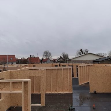 Holzrahmenbau auf Baustelle, grauer Himmel, Häuser im Hintergrund.