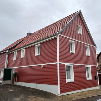 Zweistöckiges Haus mit Spitzdach welches mit fast vollständig mit rot angestrichenem Holz verkleidet ist.